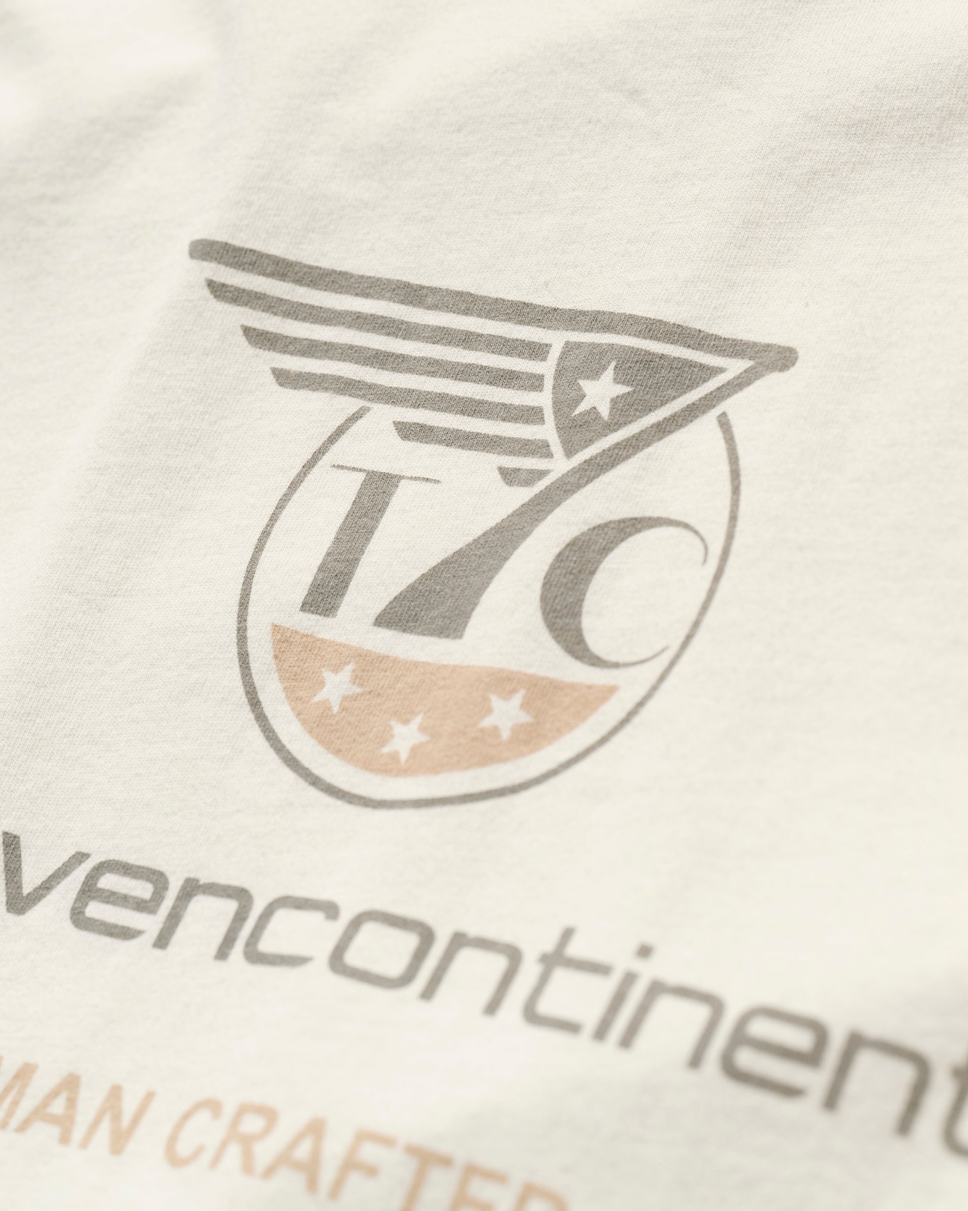 Close-up of the PRE-ORDER THE KNOXS TEE WITH STATION PRINT by T7C, made in Los Angeles with stone washed fabric, featuring a printed logo with stylized wings, 7C in a circle, stars, and faded gray and tan text.