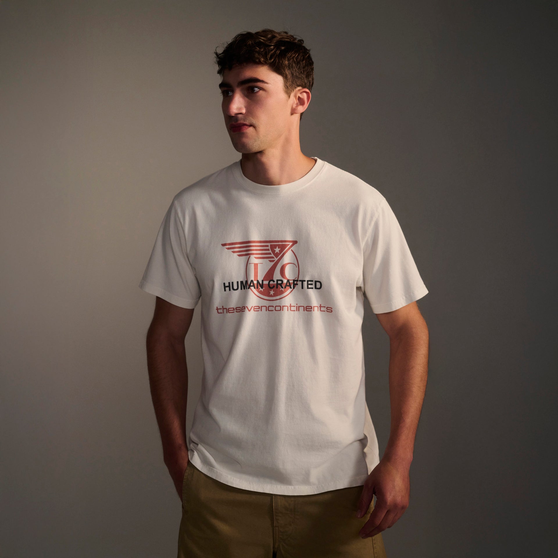 A young man with short brown hair wears tan shorts and the T7C THE KNOXS TEE WITH ICON PRINT—a white t-shirt featuring HUMAN CRAFTED thesevencontinents and a red-winged logo—against a plain background.