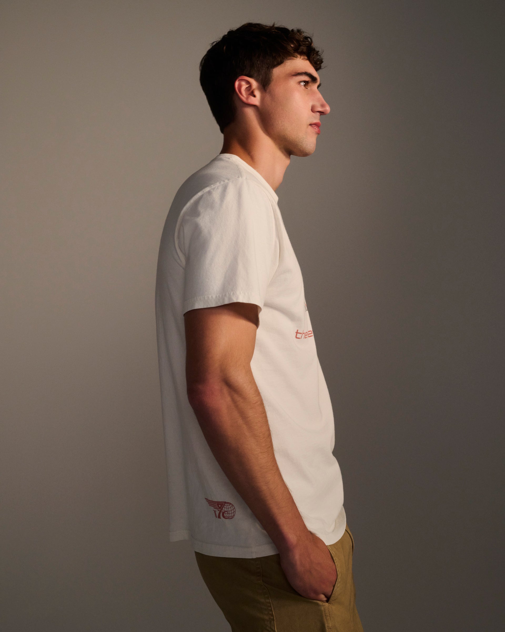 A young man with short dark hair stands in profile against a neutral background, wearing the T7C THE KNOXS TEE WITH ICON PRINT in white and tan pants, his hands tucked into his pockets.