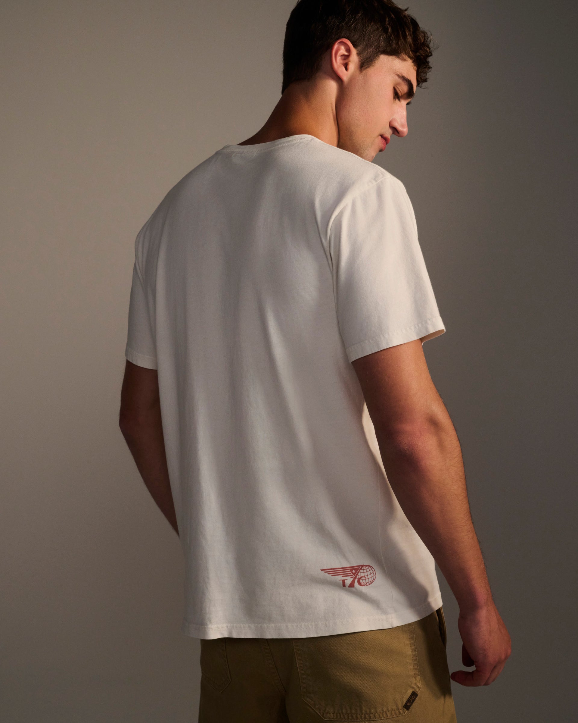 A young man faces away from the camera in a T7C THE KNOXS TEE WITH ICON PRINT—a plain white t-shirt with a small red graphic near the hem—paired with khaki shorts against a neutral background.