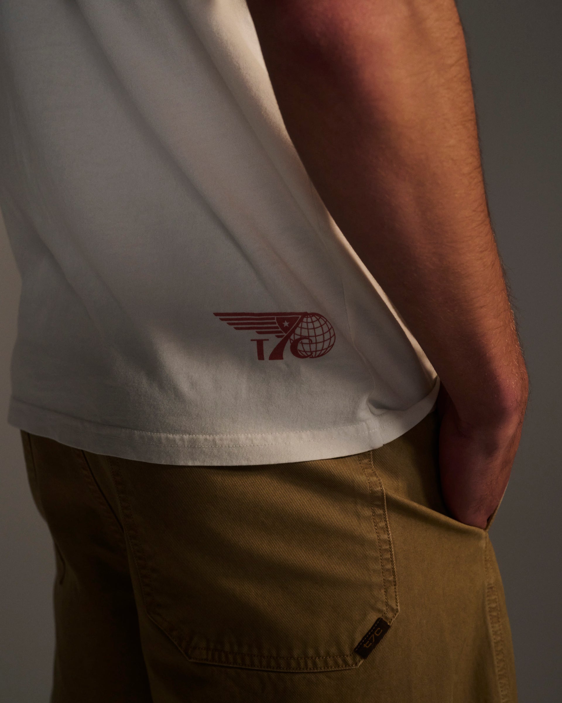 A person wears the T7C THE KNOXS TEE WITH ICON PRINT, a white shirt with a red graphic, paired with tan pants; only their torso and upper legs are shown, with one hand in their pocket.