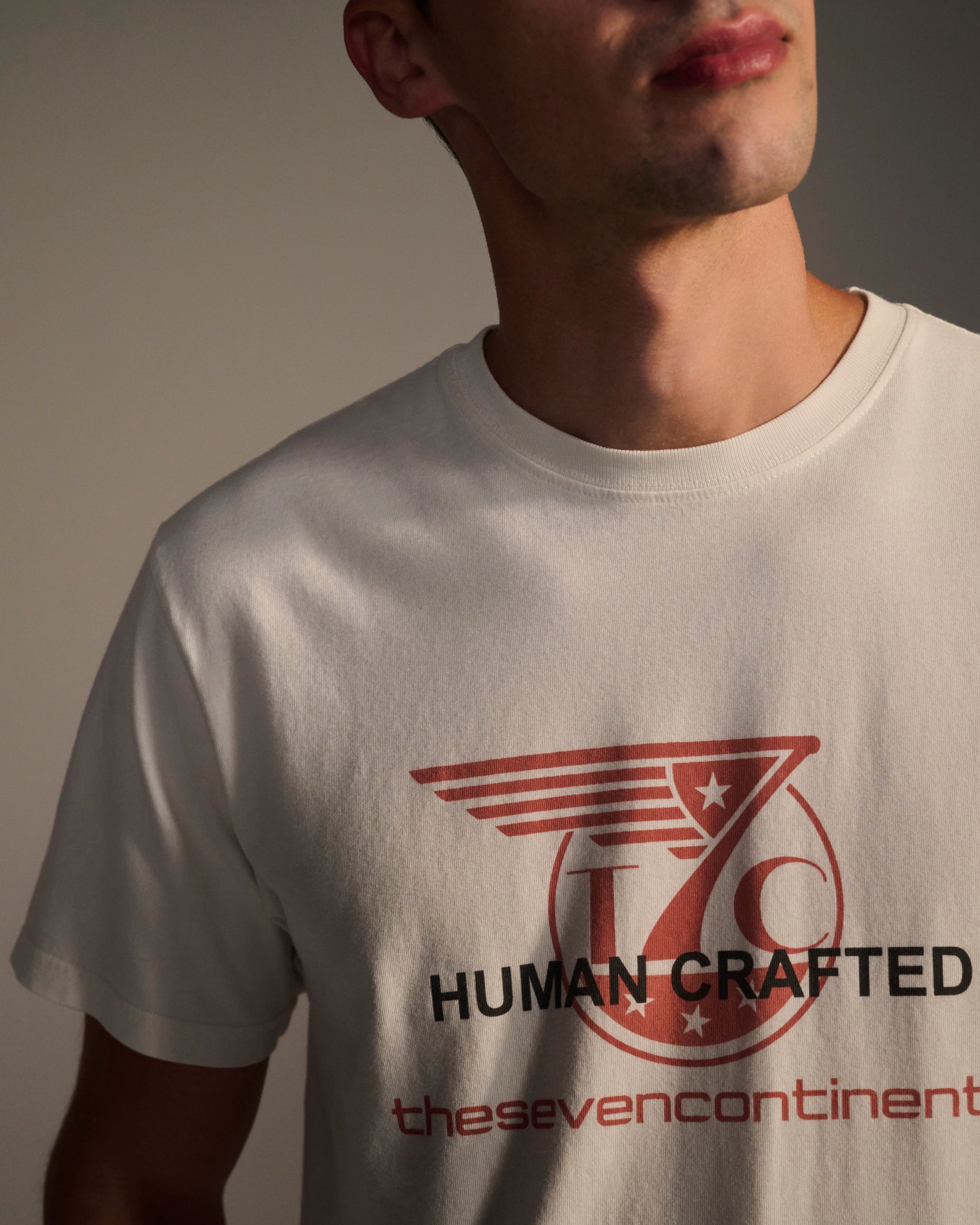 A person wearing the T7C THE KNOXS TEE WITH ICON PRINT—a white shirt with a red and black winged star logo and the text HUMAN CRAFTED and thesevencontinents. The wearer’s face is partially out of frame.