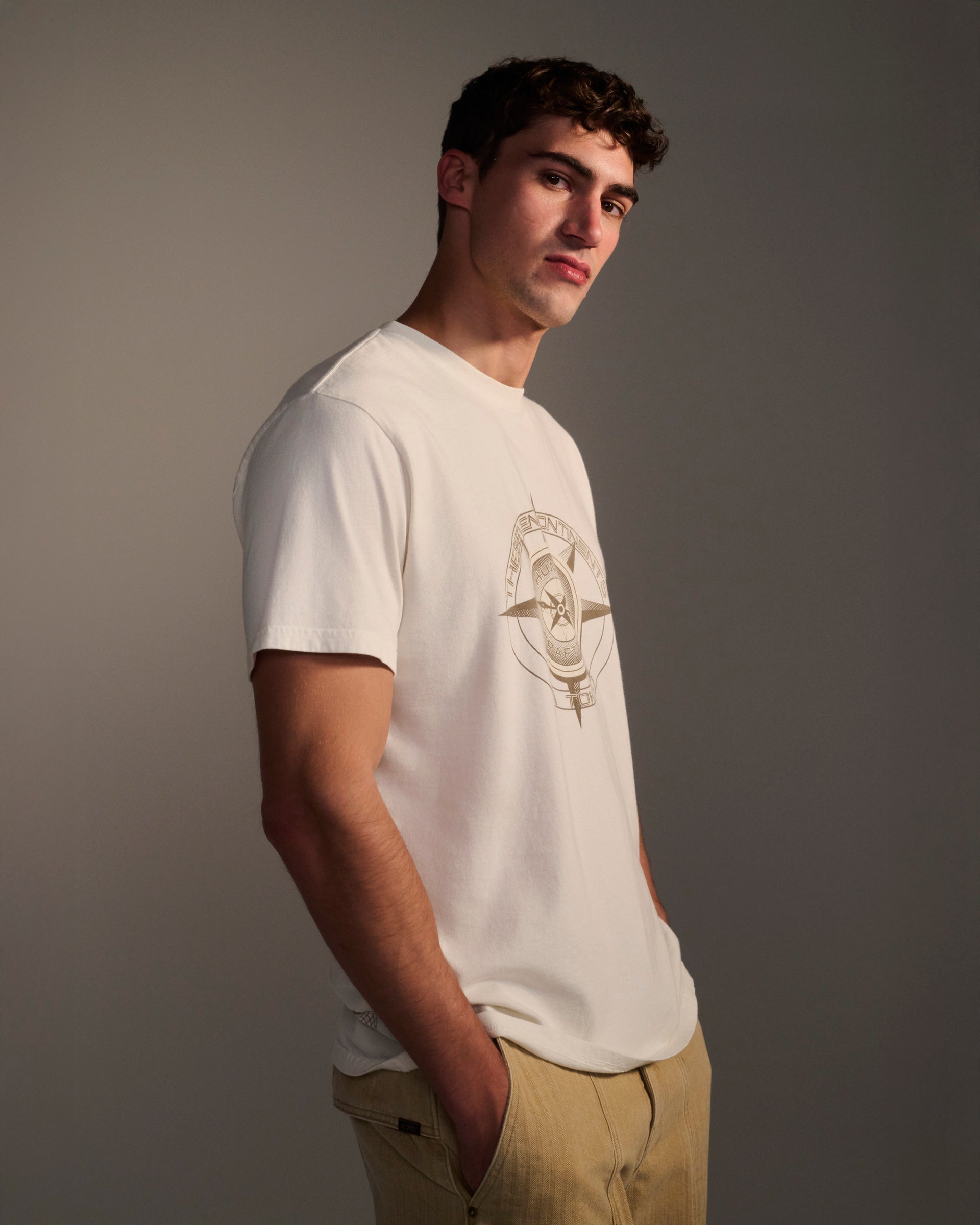 A young man with short curly hair stands hands in pockets, wearing the T7C PRE-ORDER: THE KNOXS TEE WITH MERIDIAN PRINT and light beige pants, looking slightly to the side against a plain background.