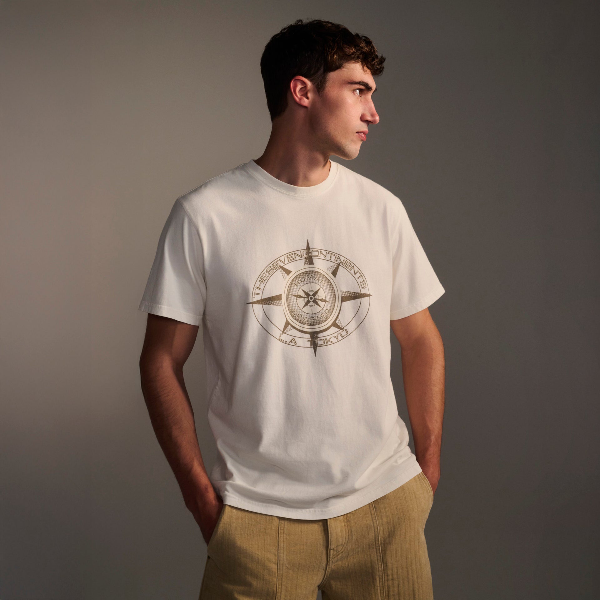 A young man wears the T7C PRE-ORDER: THE KNOXS TEE WITH MERIDIAN PRINT, featuring an eye graphic and text, with his hands in tan pants pockets, looking to the side against a neutral background.