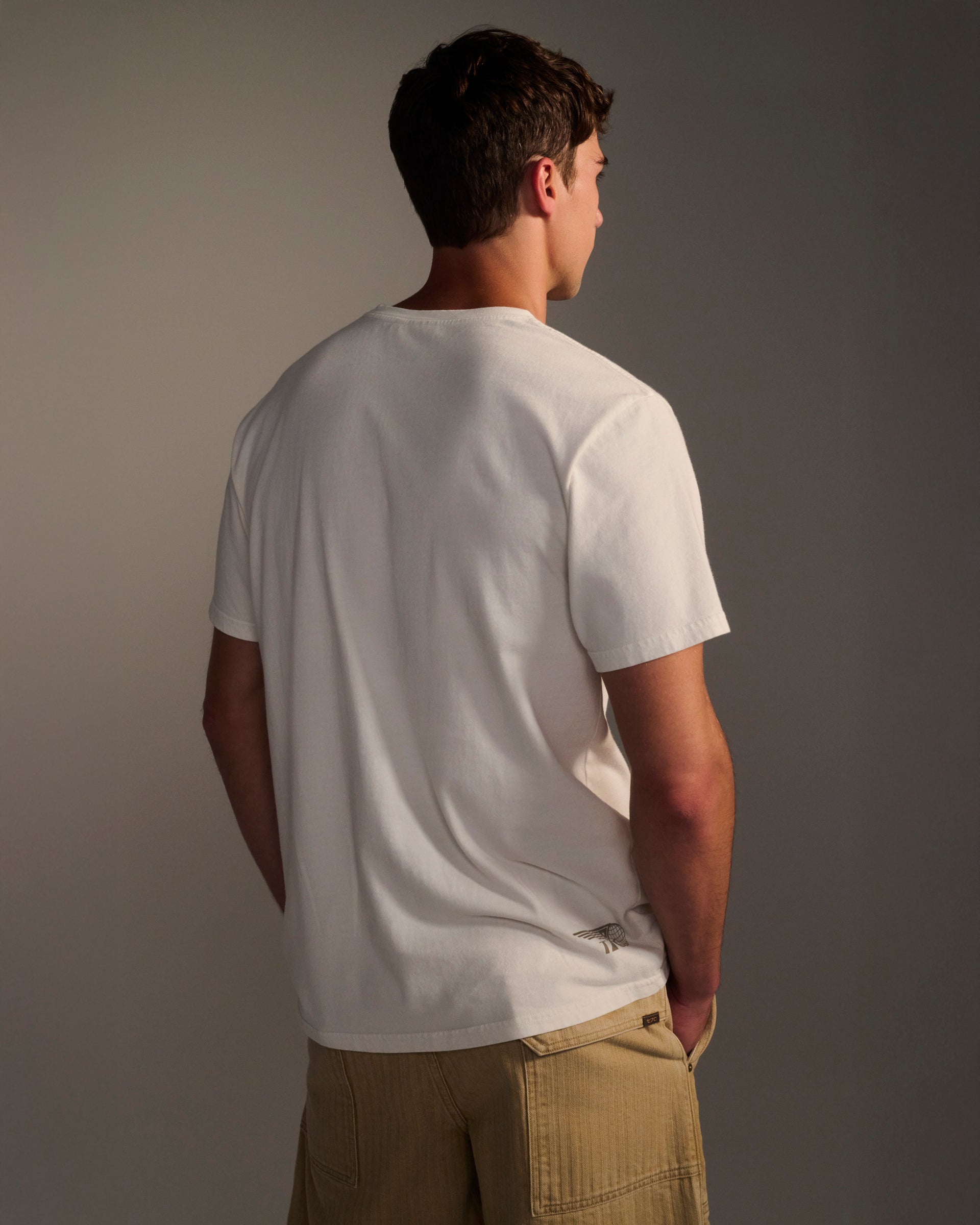 A man stands with his back to the camera, wearing the T7C PRE-ORDER: THE KNOXS TEE WITH MERIDIAN PRINT and light brown shorts against a neutral background, arms relaxed by his sides.