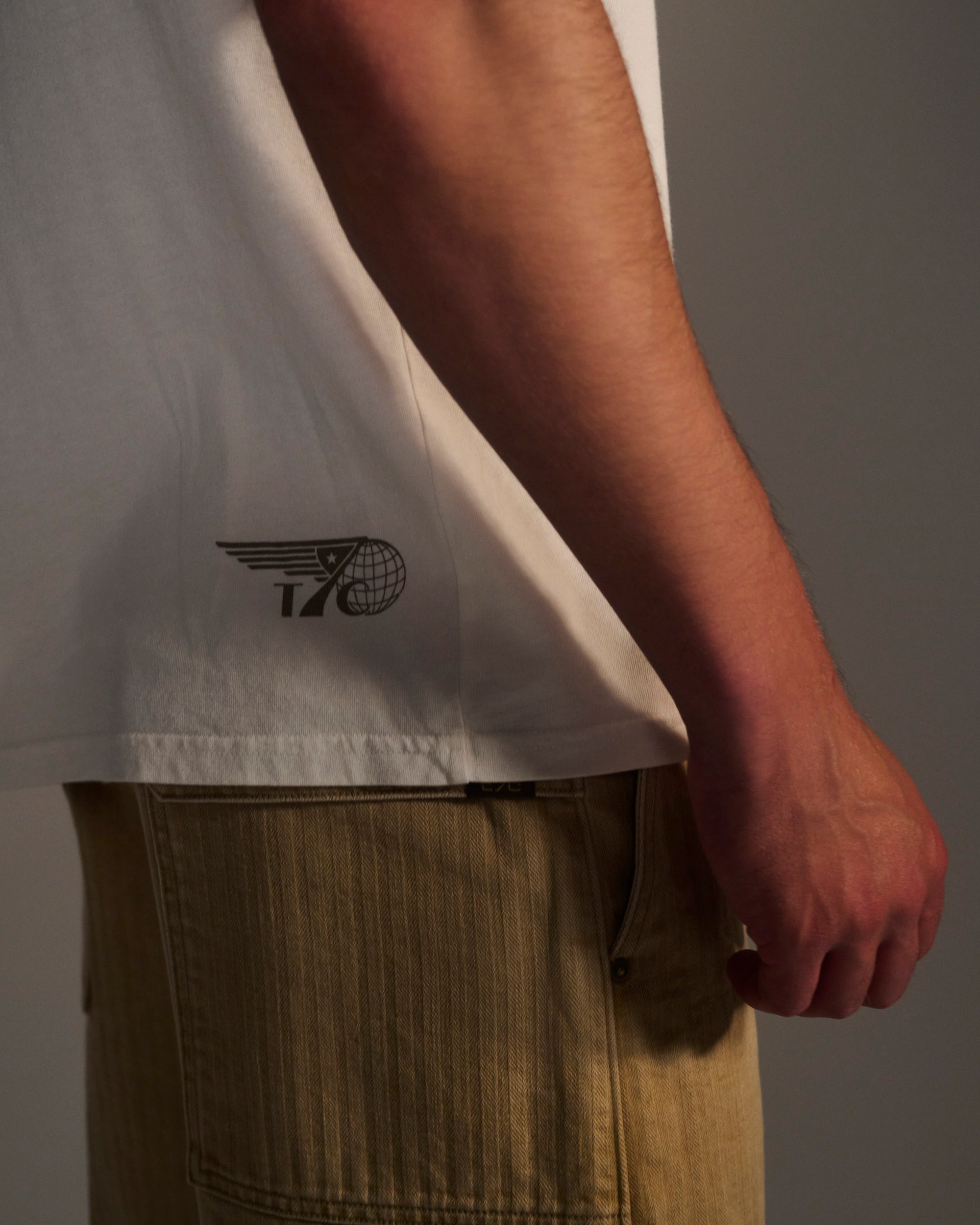 A person is shown from the side wearing the T7C PRE-ORDER: THE KNOXS TEE WITH MERIDIAN PRINT, featuring a winged globe logo, paired with tan pants and their arm relaxed by their waist.