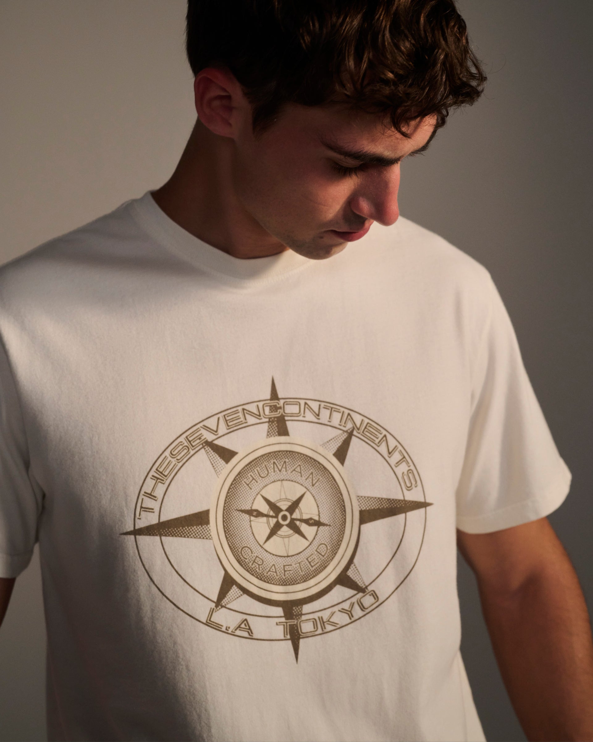 A young man wears the T7C PRE-ORDER: THE KNOXS TEE WITH MERIDIAN PRINT, featuring a compass and THE SEVEN CONTINENTS, HUMAN CRAFTED, L.A. TOKYO on the front. He looks down against a plain background.