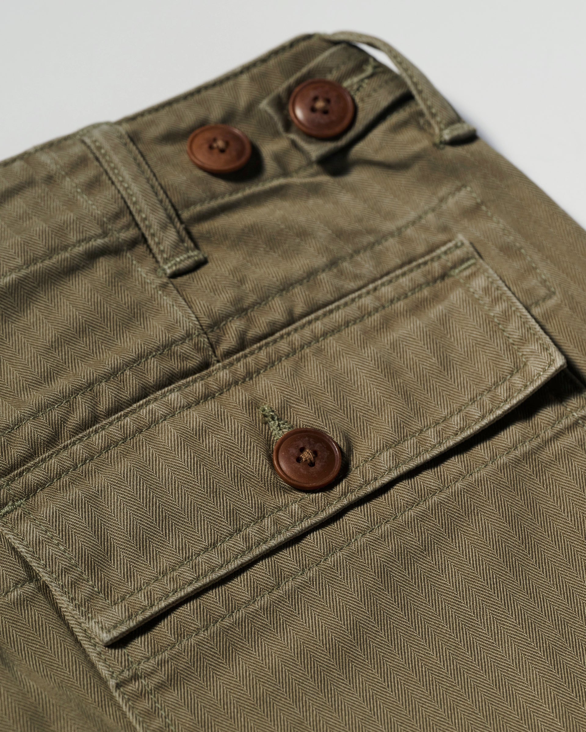 The T7C Liam Military Pant in Olive Drab features cotton herringbone fabric, brown buttons, a belt loop, a flap pocket with visible stitching, and a relaxed-fit design inspired by classic military styles.