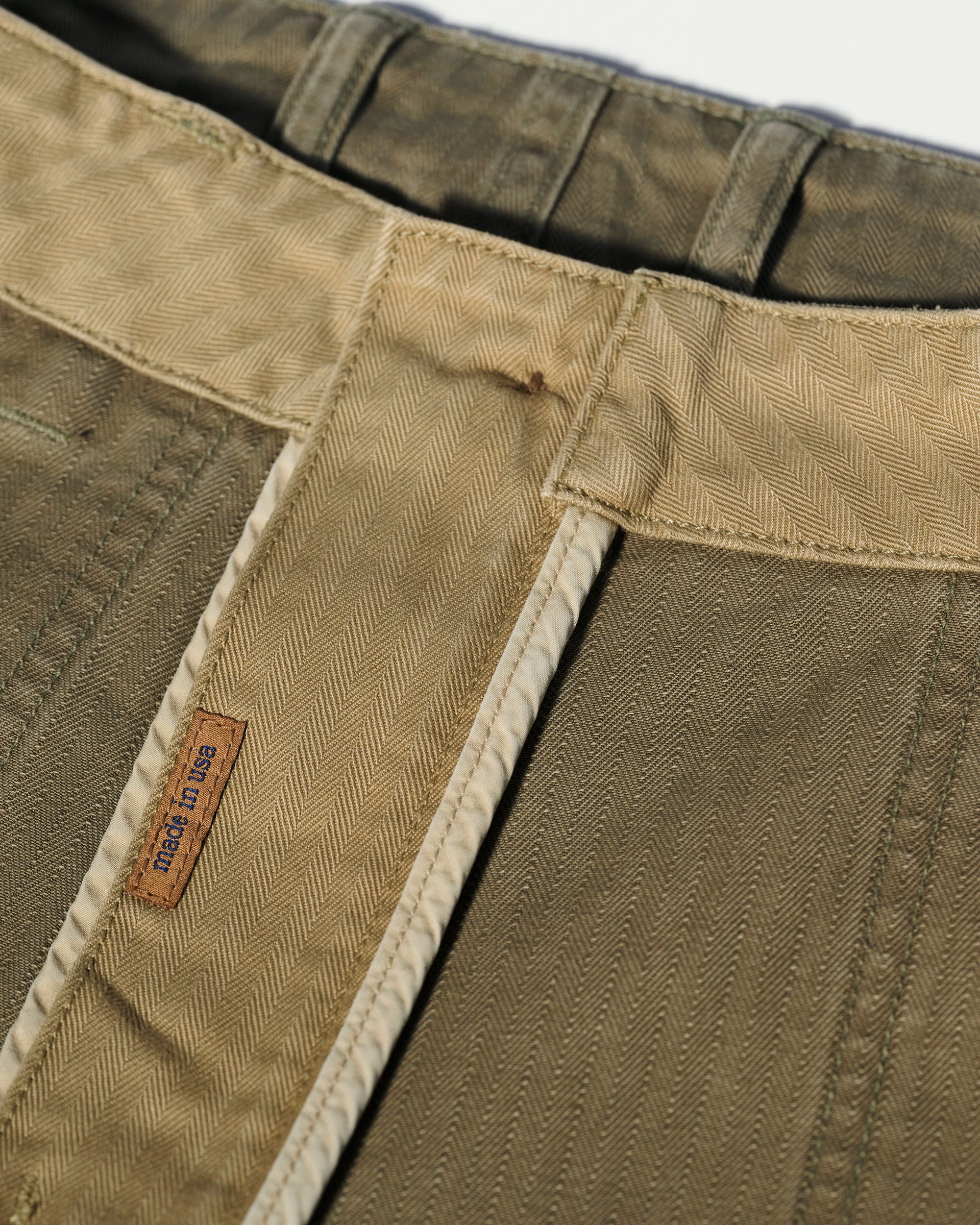 Close-up of the waistband and inner stitching of The Liam Military Pant in Olive Drab by T7C, crafted from cotton herringbone fabric with detailed patterns, belt loops, and a small brown label featuring blue Sun House text.