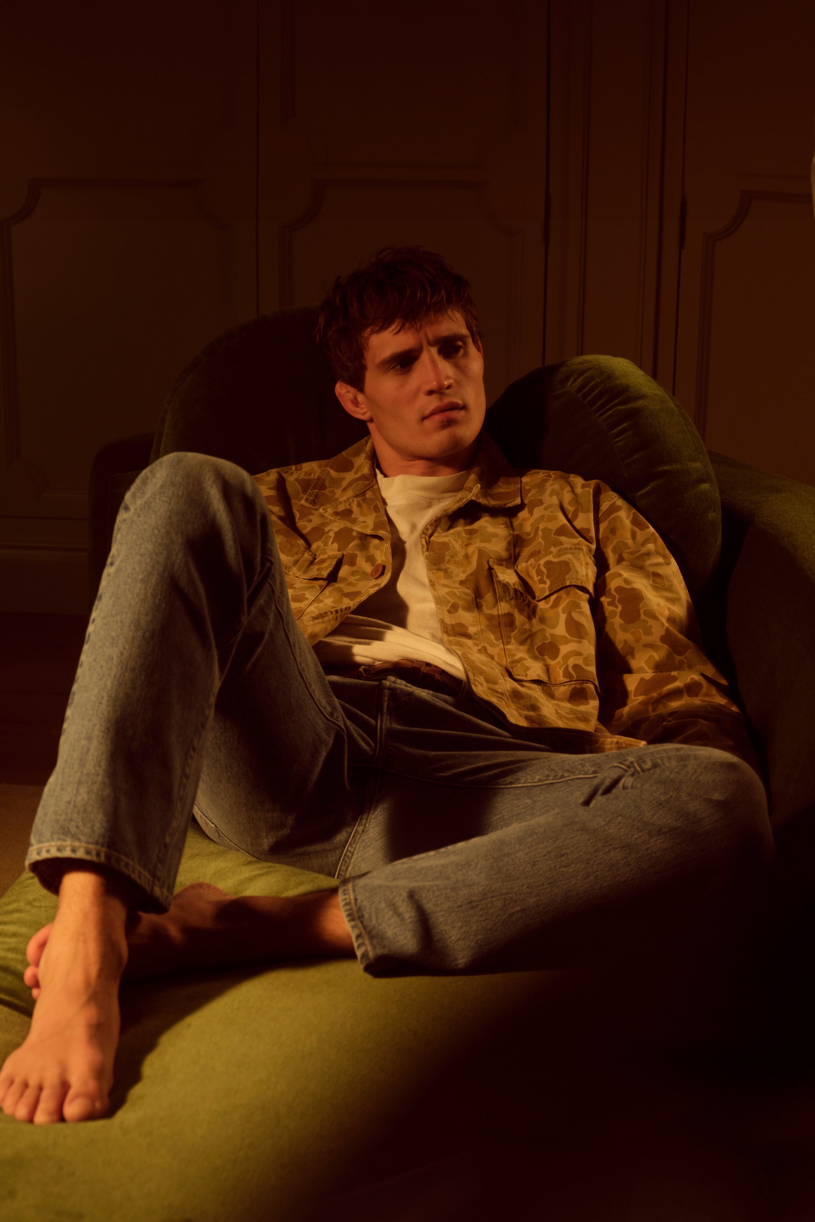 A male model in jeans and a camouflage jacket sitting on a green couch in a dark room