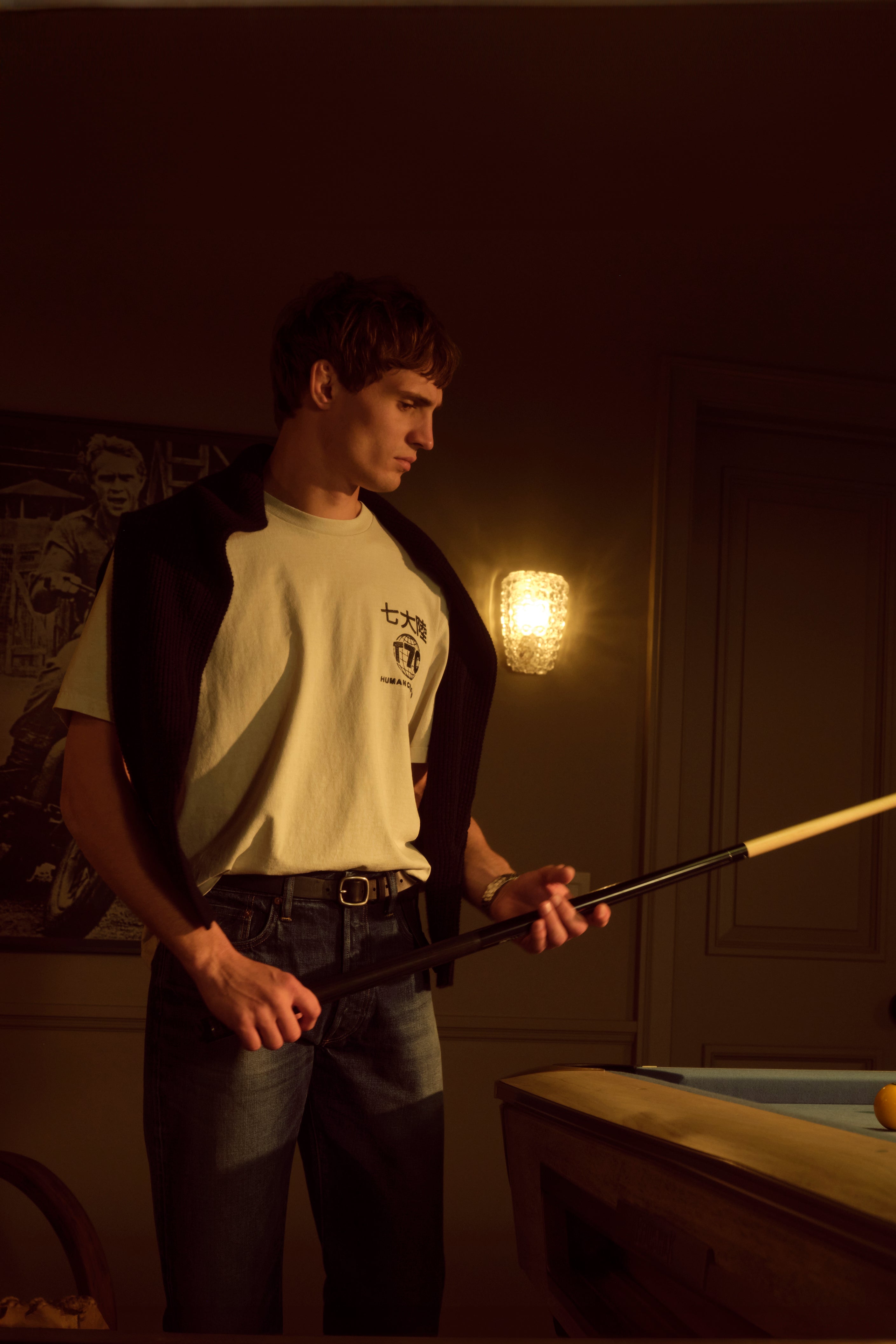 A male model wearing a t-shirt and jeans playing pool in a dark room