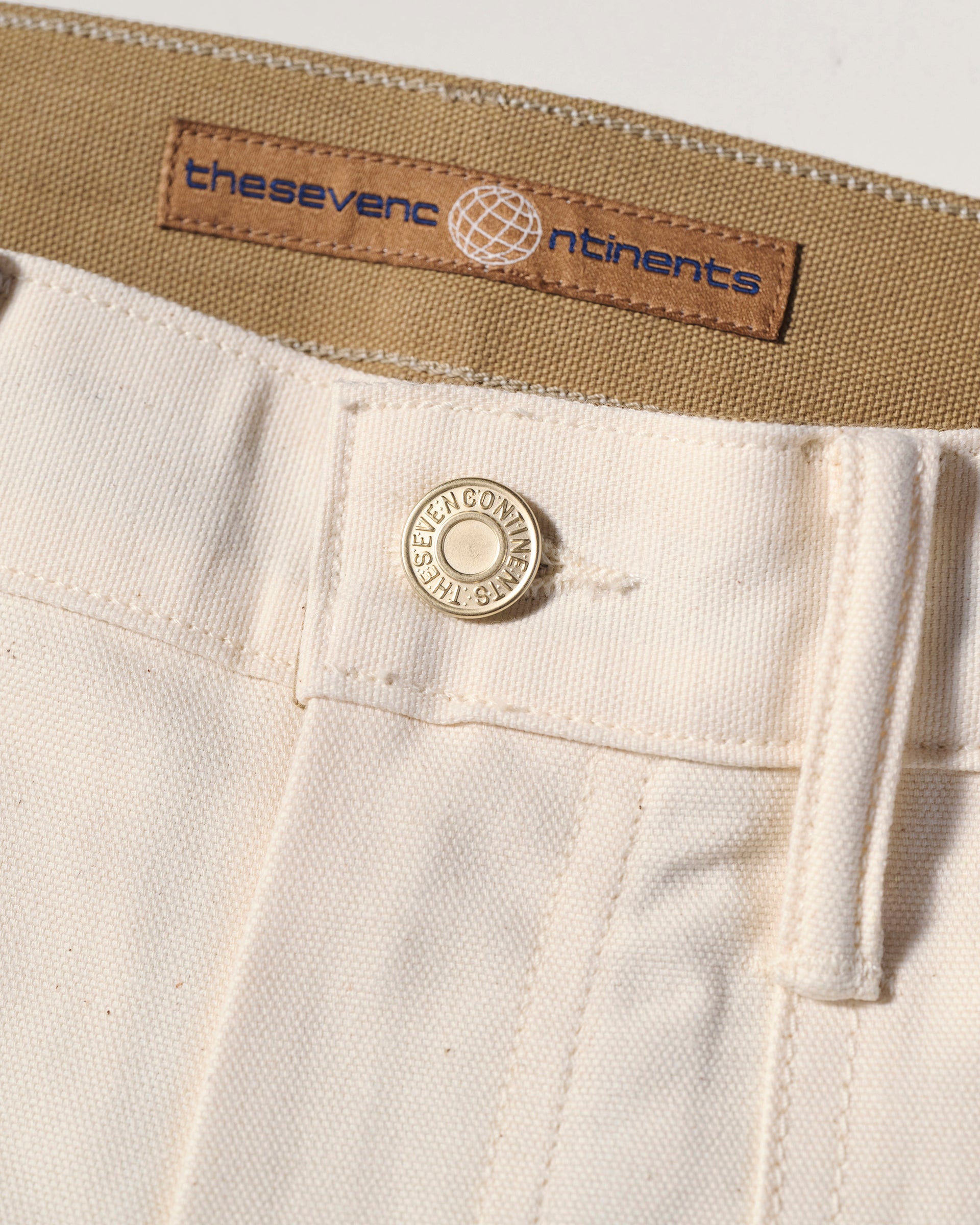 Close-up of The Sid Pant in Chalk Raw by T7C The Seven Continents, featuring a metal button engraved with THE SEVEN CONTINENTS and a tan label with brand name and globe logo above the zipper, inspired by classic workwear.