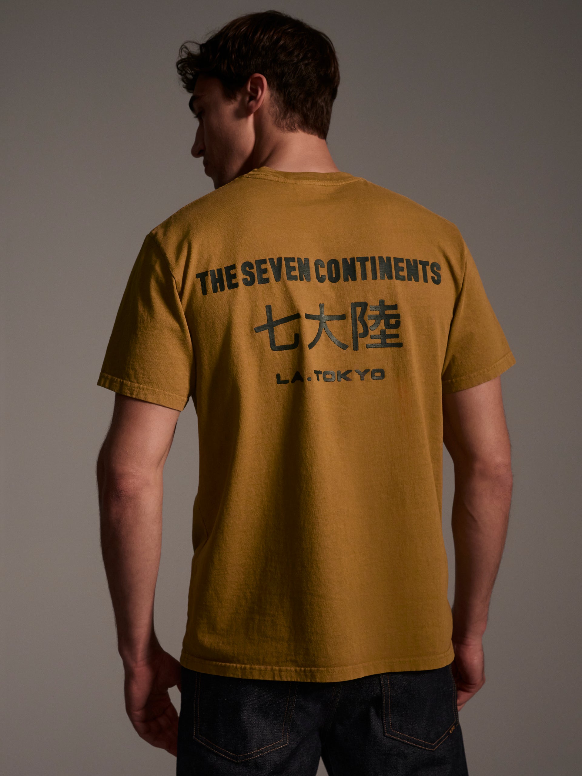 A back image of a male model wearing a mustard t-shirt with a logo detail