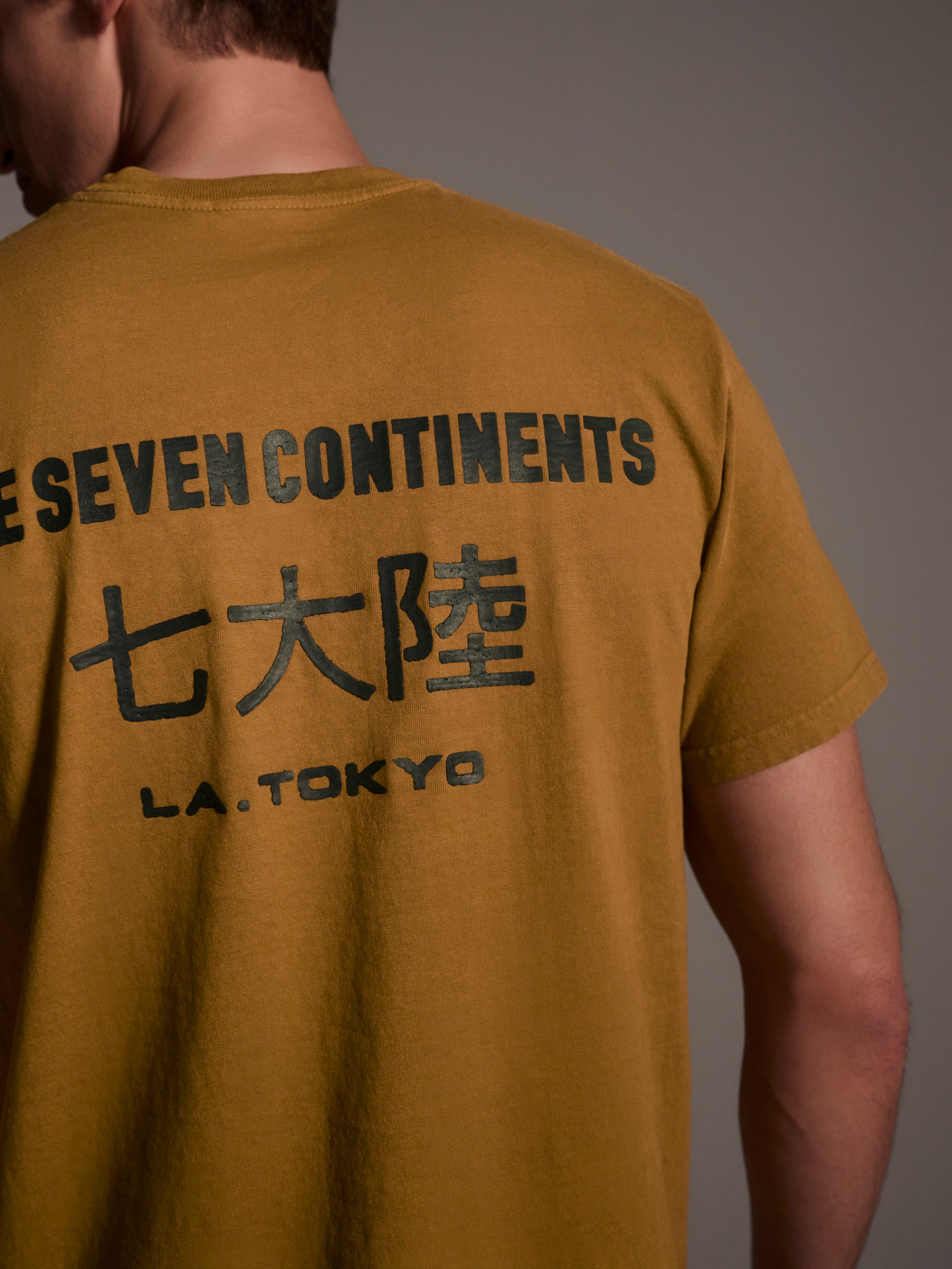 A back detail image of a male model wearing a mustard t-shirt with a logo detail