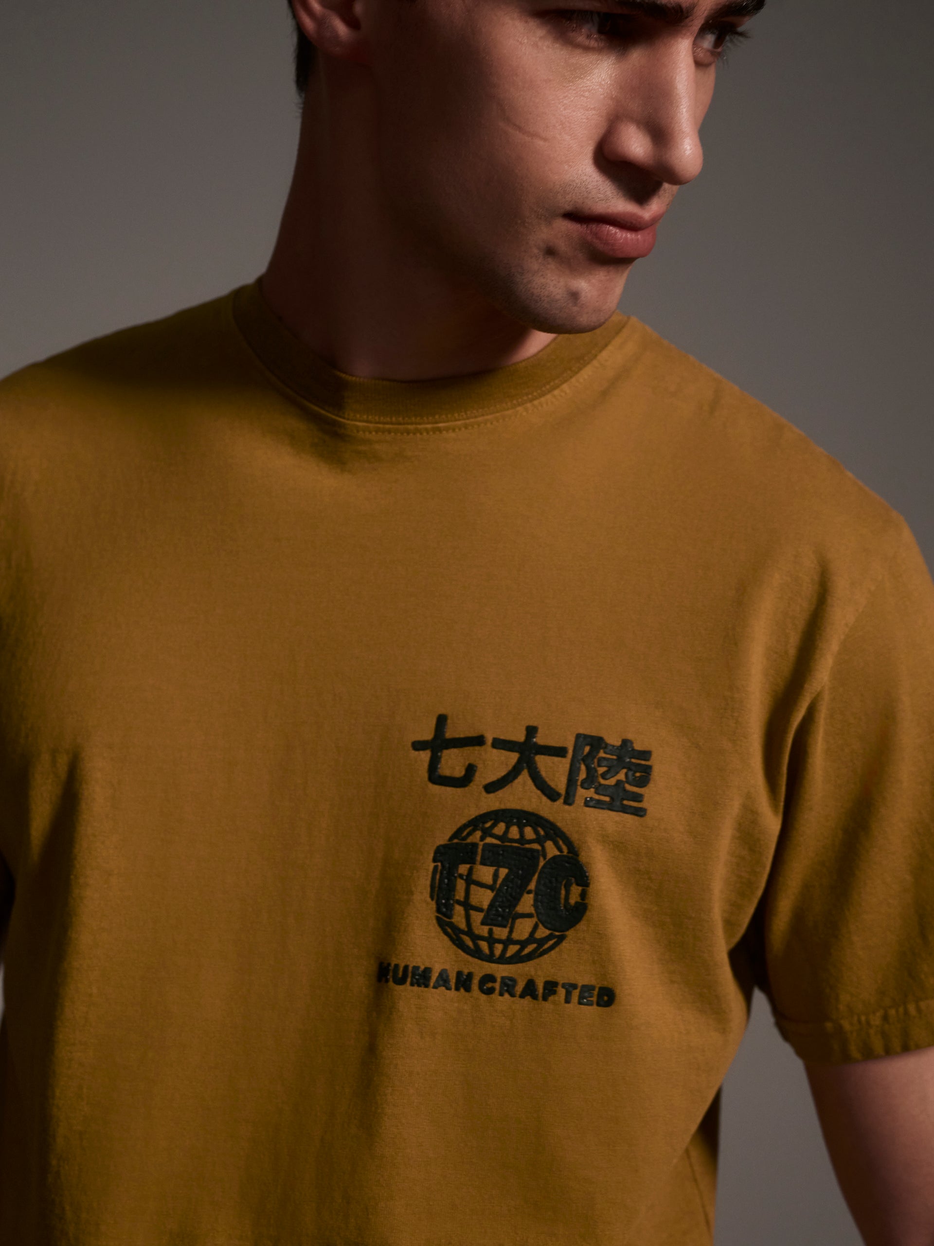 A detail image of a male model wearing a mustard t-shirt with a logo detail