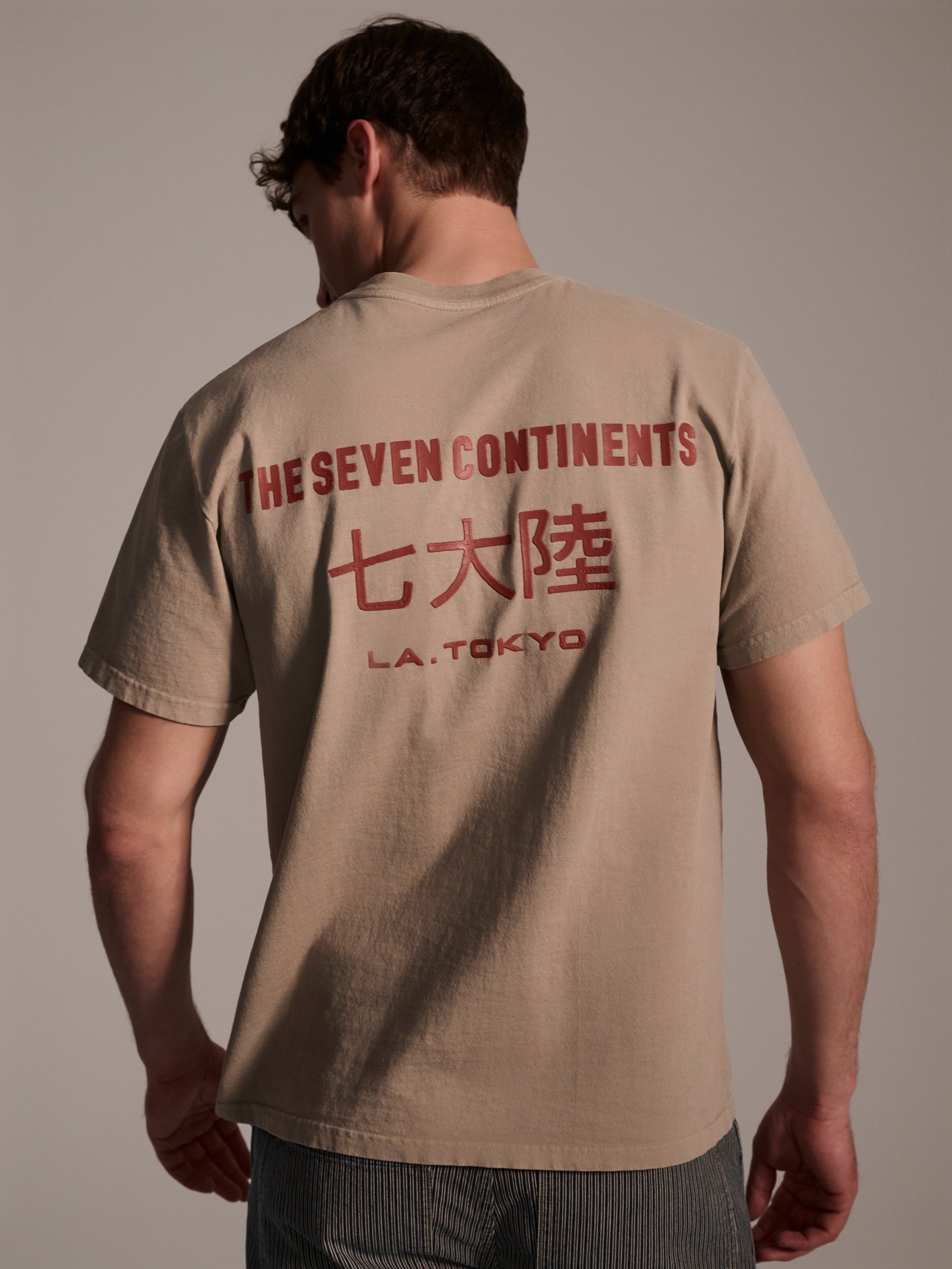 A back image of a male model in a beige t-shirt with a red logo detail