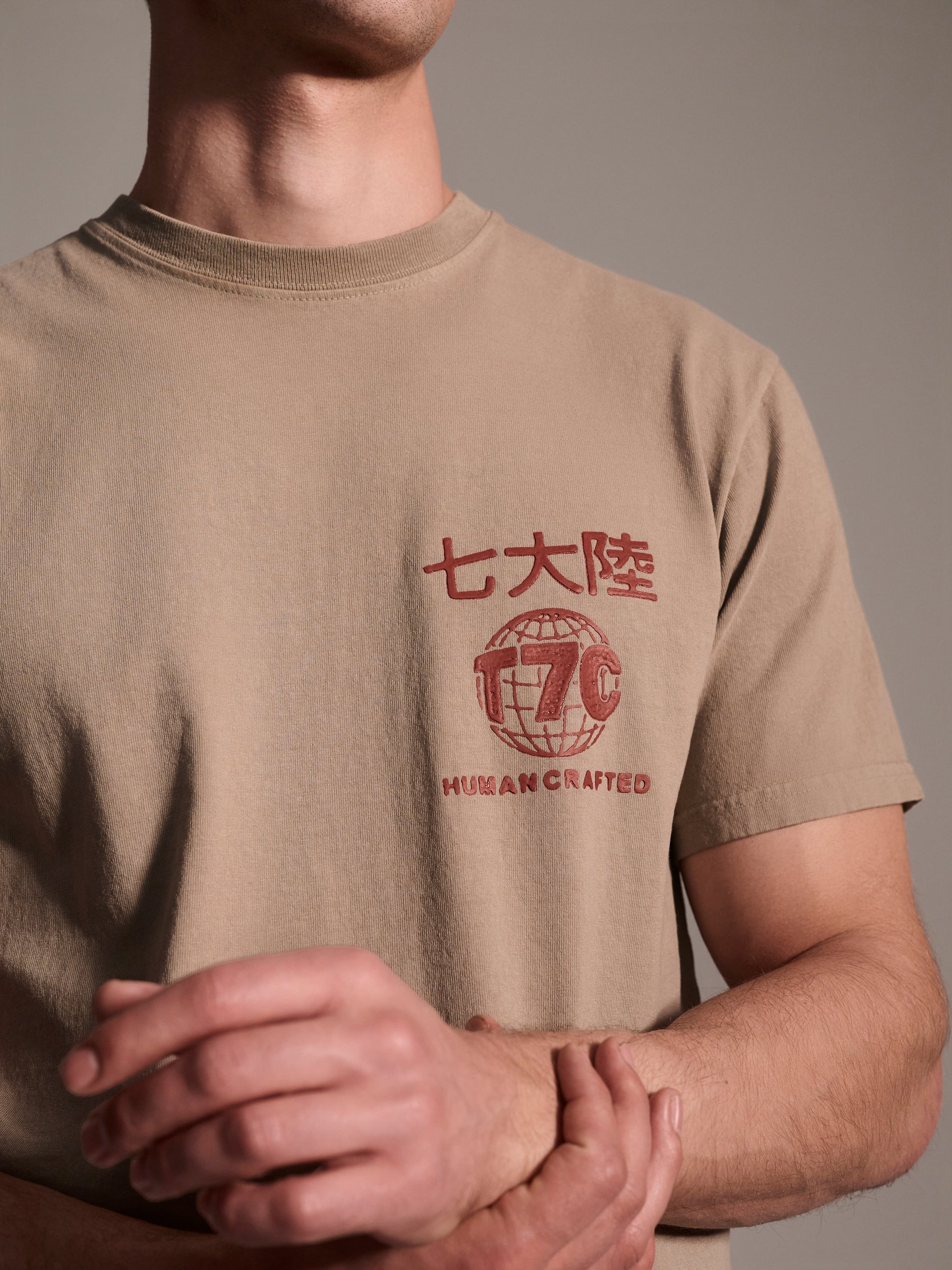 A detail image of a male model in a beige t-shirt with a red logo detail