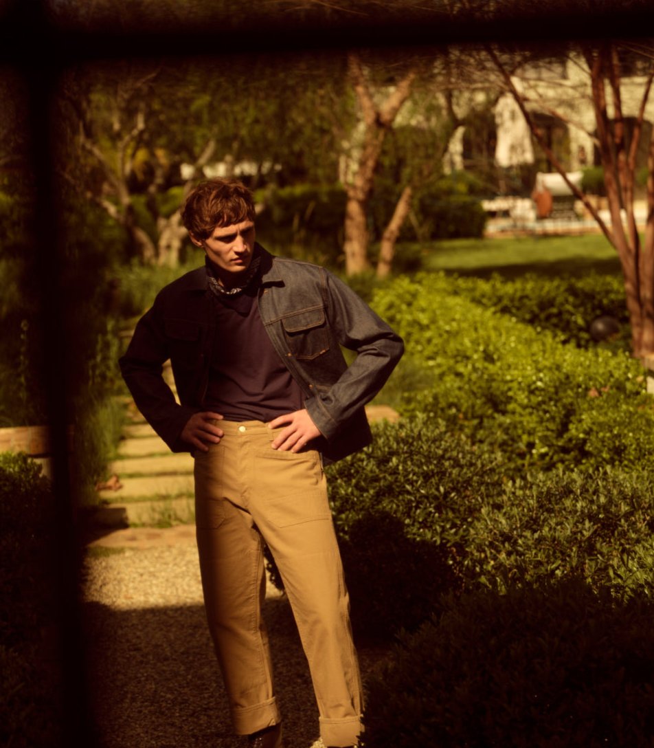 A male model wearing a denim jacket and khaki pants in a garden