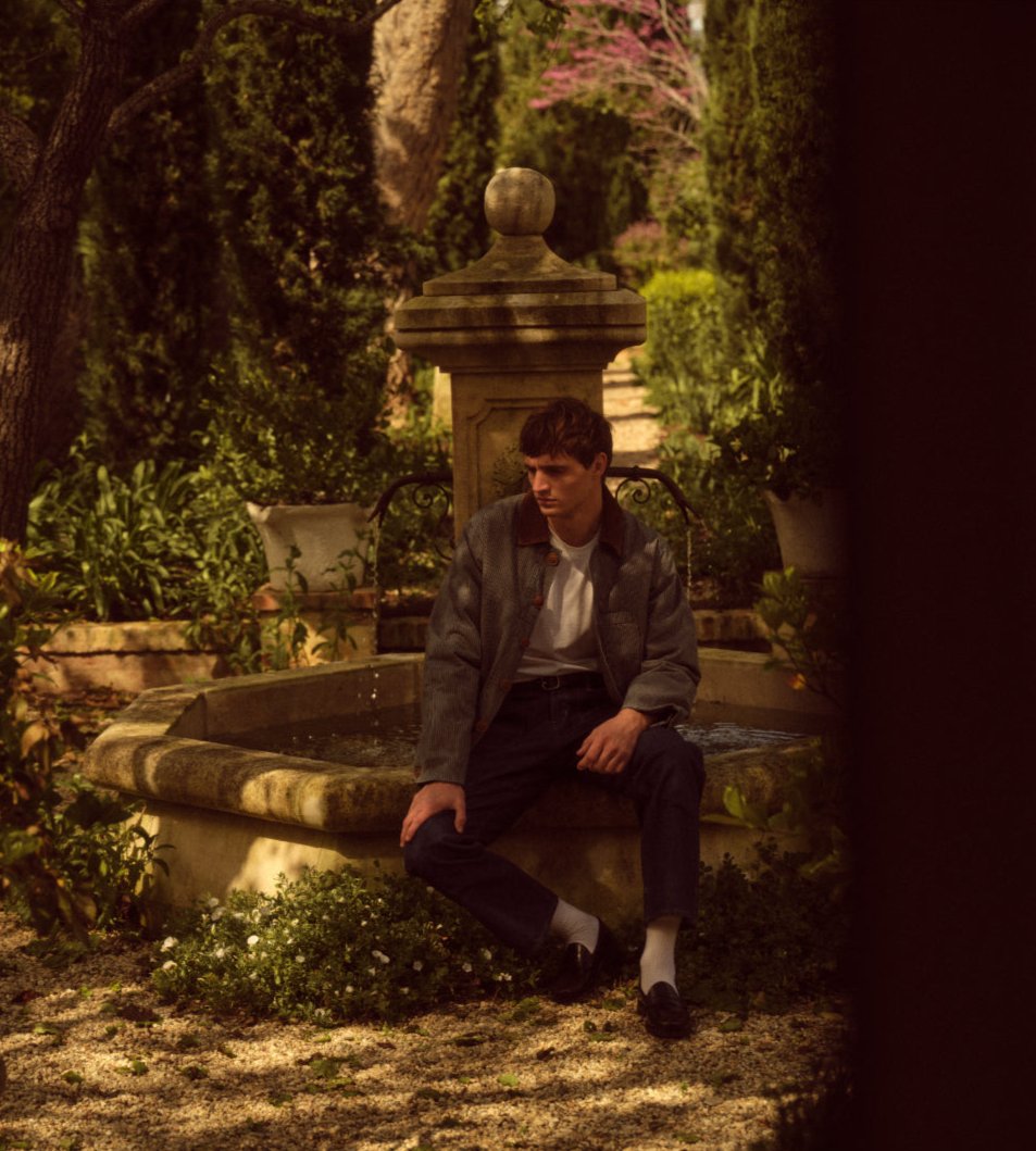 A male model wearing a striped jacket and jeans sitting on a fountain in a garden