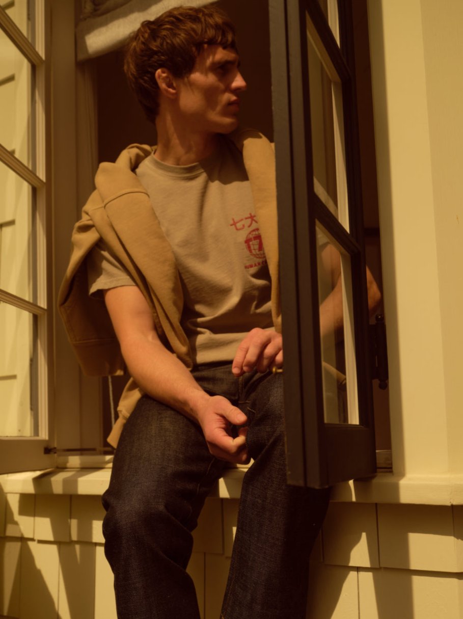 A male model wearing a t-shirt and jeans sitting on a window sill