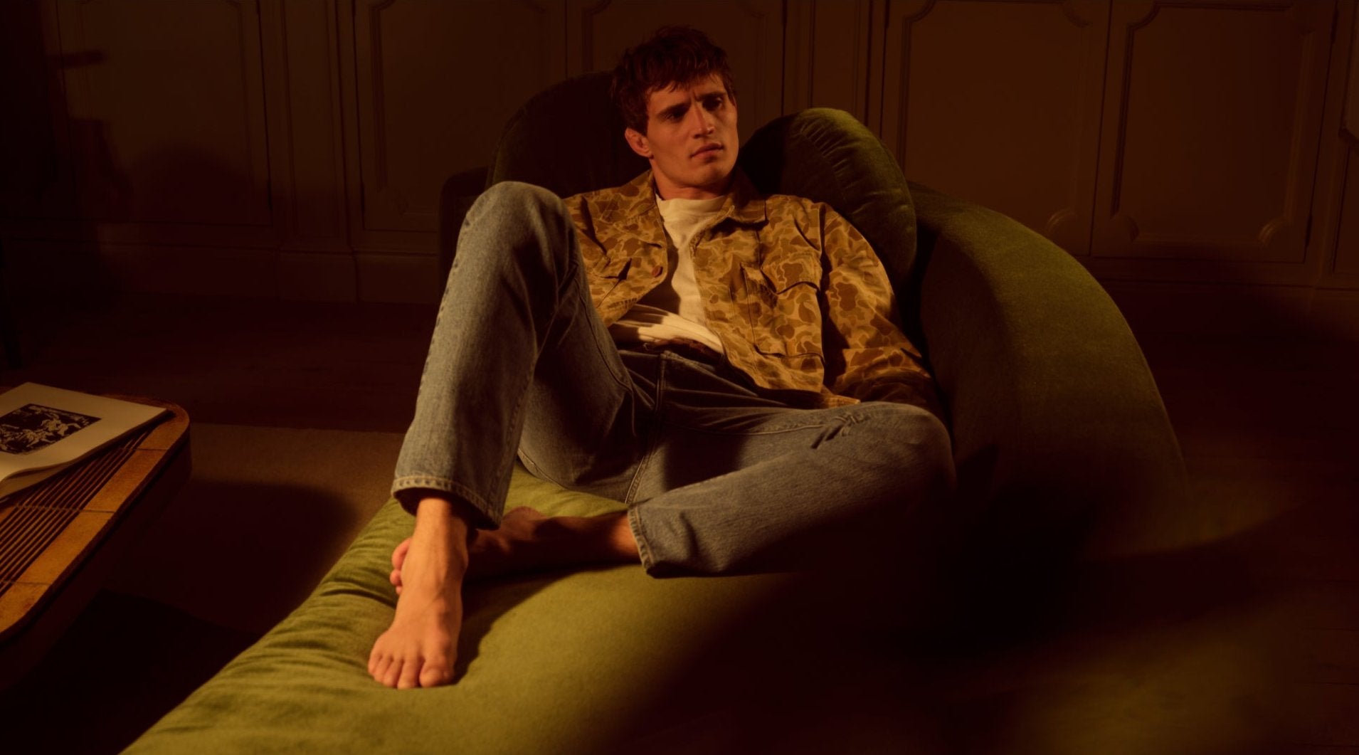 A male model in jeans and a camouflage jacket sitting on a green couch in a dark room