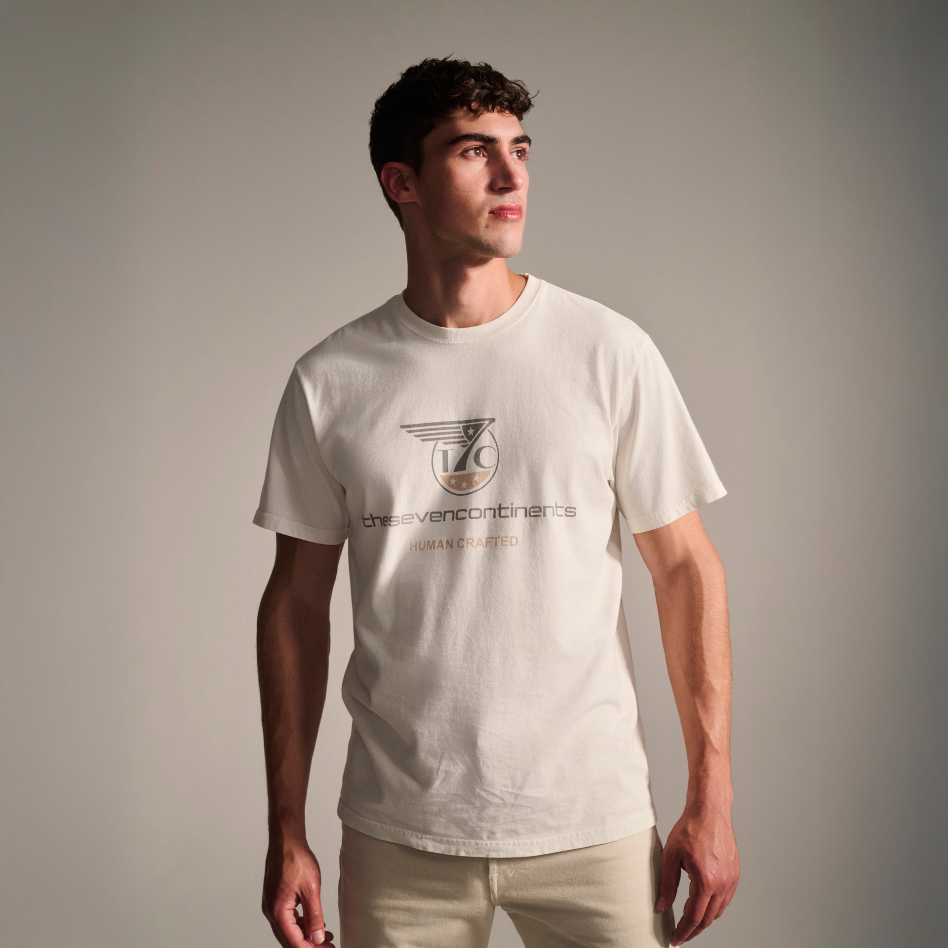 A young man stands against a plain background, wearing the T7C PRE-ORDER THE KNOXS TEE WITH STATION PRINT—a stone-washed, 100% cotton white T-shirt featuring the seven continents and HUMAN CRAFTED on the front.
