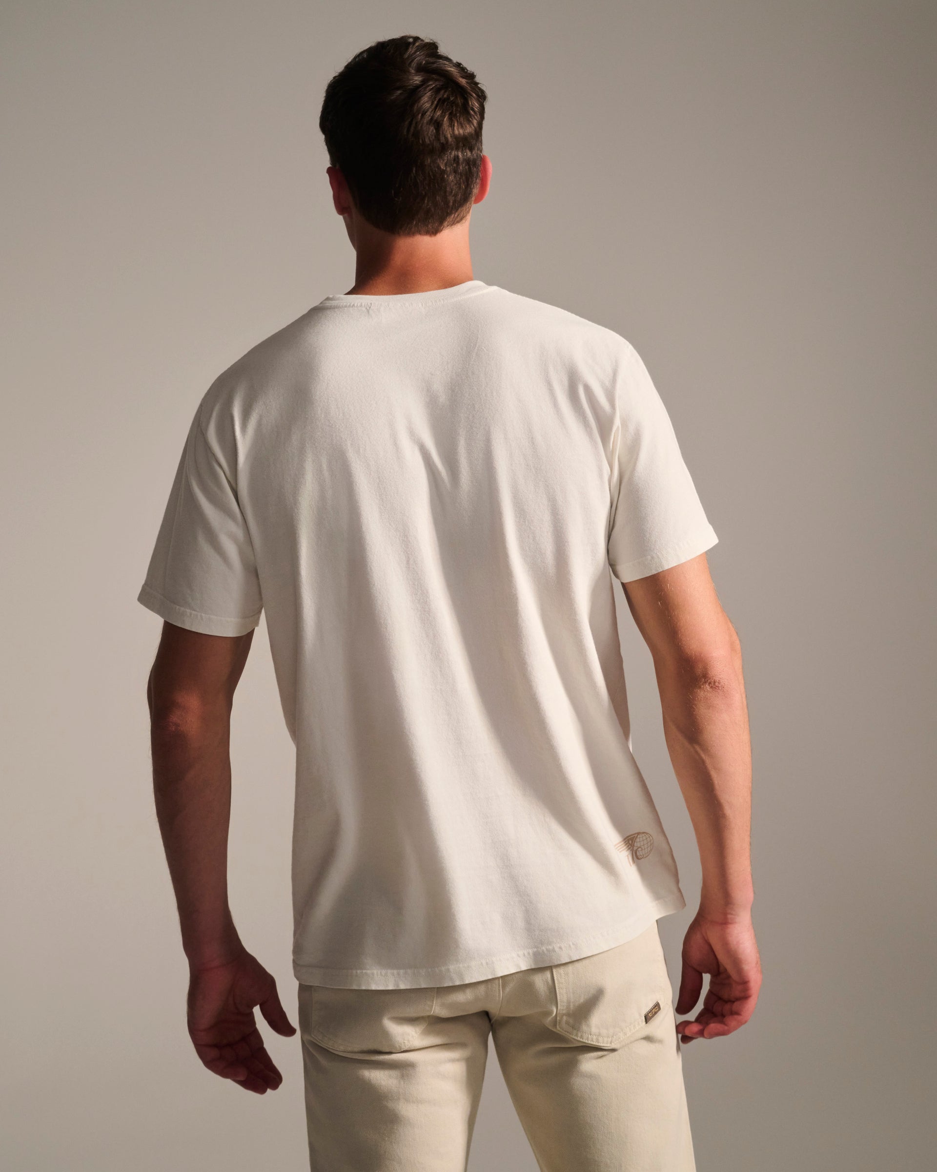 A man faces away, wearing the T7C PRE-ORDER THE KNOXS TEE WITH STATION PRINT and light beige pants against a neutral background.