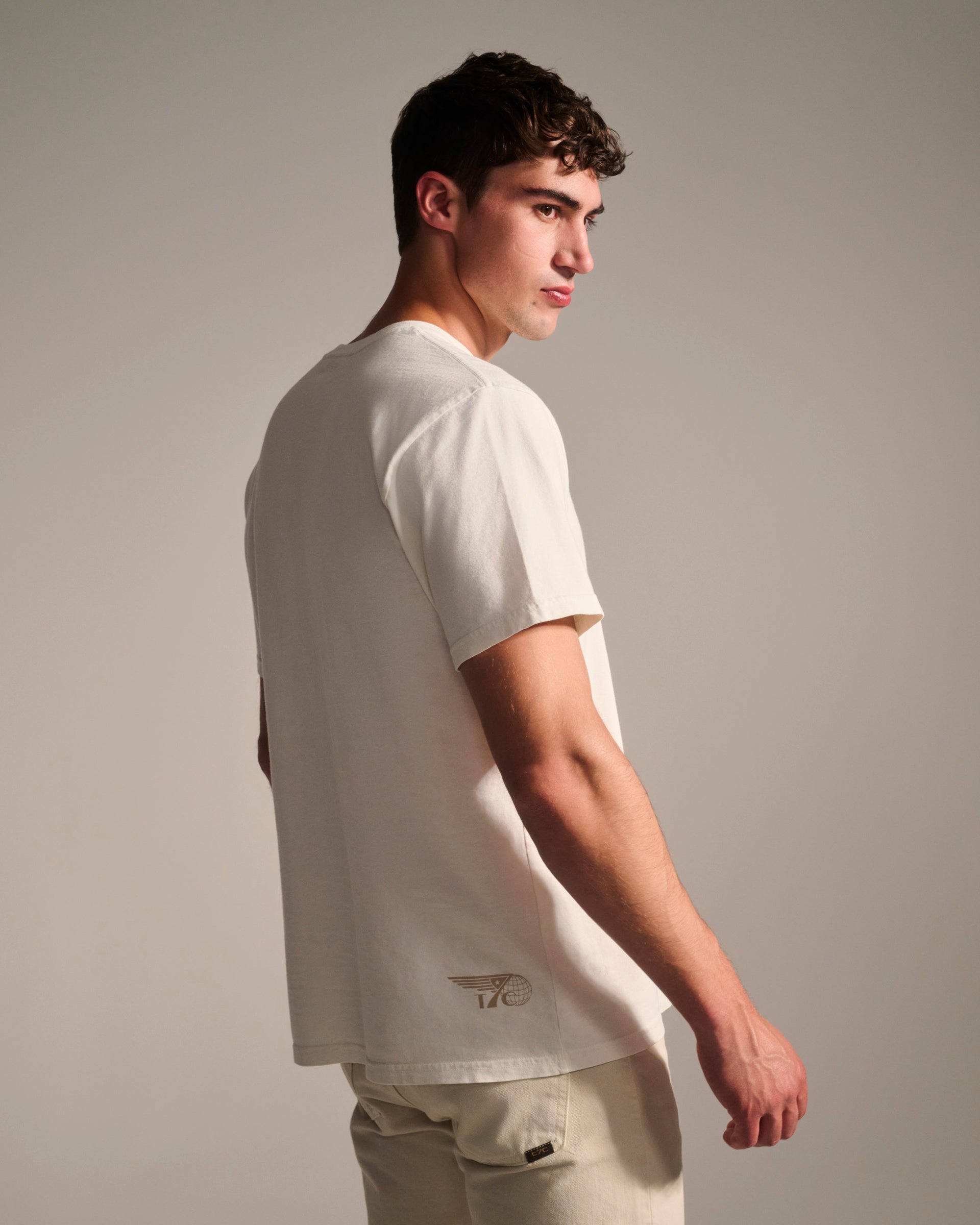 A young man with short brown hair models the T7C PRE-ORDER THE KNOXS TEE WITH STATION PRINT, a stone washed 100% cotton shirt featuring a small back graphic near the hem, standing sideways against a plain background.