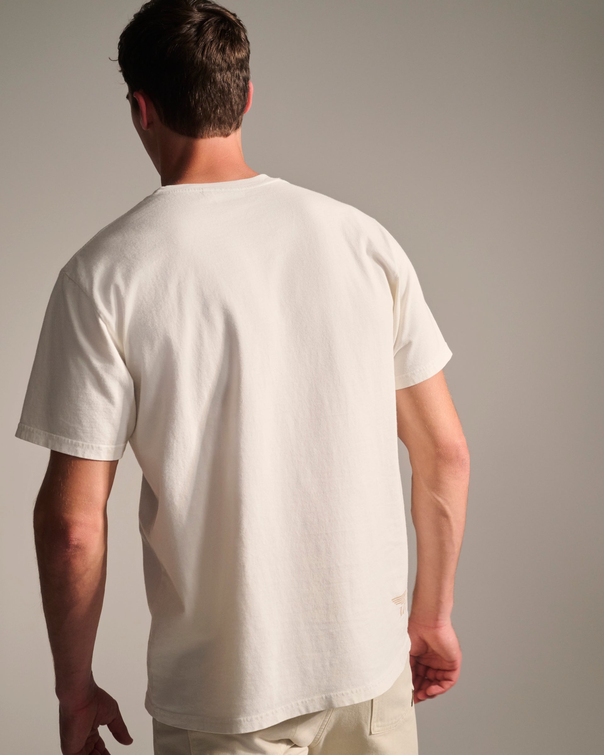A man with short brown hair, seen from the back against a neutral background, wears light pants and the T7C PRE-ORDER THE KNOXS TEE WITH STATION PRINT.