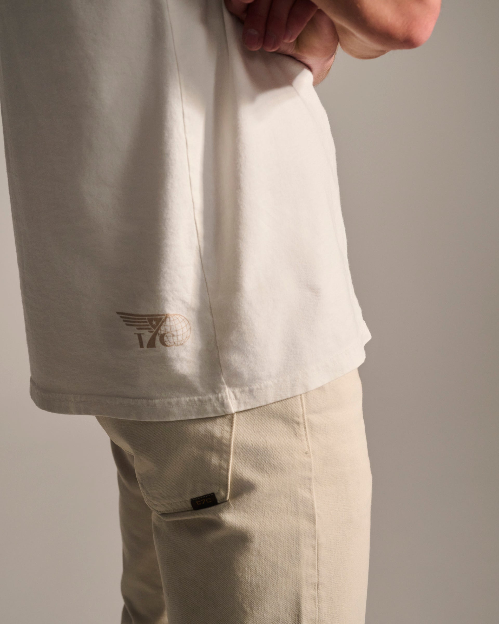 Wearing the T7C PRE-ORDER THE KNOXS TEE WITH STATION PRINT, a person with light beige pants stands with one hand on their hip against a plain background. The tee is stone washed white and features a small tan logo near the hem.