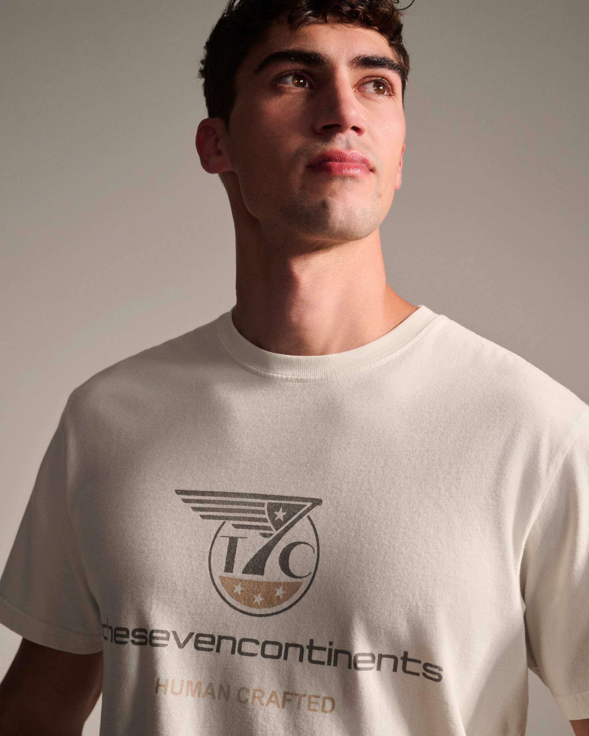 A man in a white, stone-washed T7C PRE-ORDER THE KNOXS TEE WITH STATION PRINT—featuring “thesevencontinents” and “HUMAN CRAFTED” logos—looks up neutrally against a plain background. Made in Los Angeles from 100% cotton.