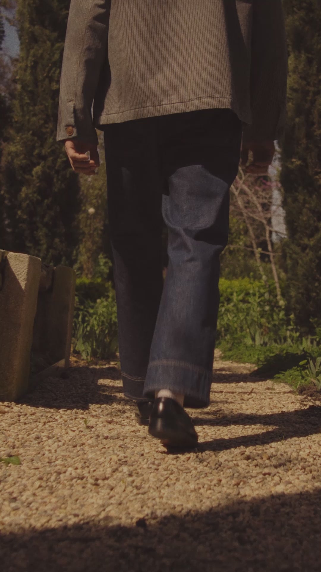 A male model wearing a railroad striped jacket and relaxed denim jeans walking in a garden